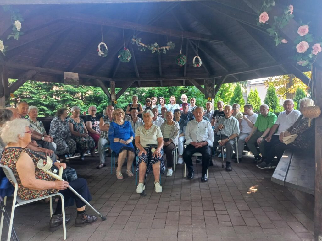 Na zdjęciu widzimy na pierwszym planie siedzących w altanie mieszkańców Domu Pomocy Społecznej, za nimi stojąca kadra z tego domu